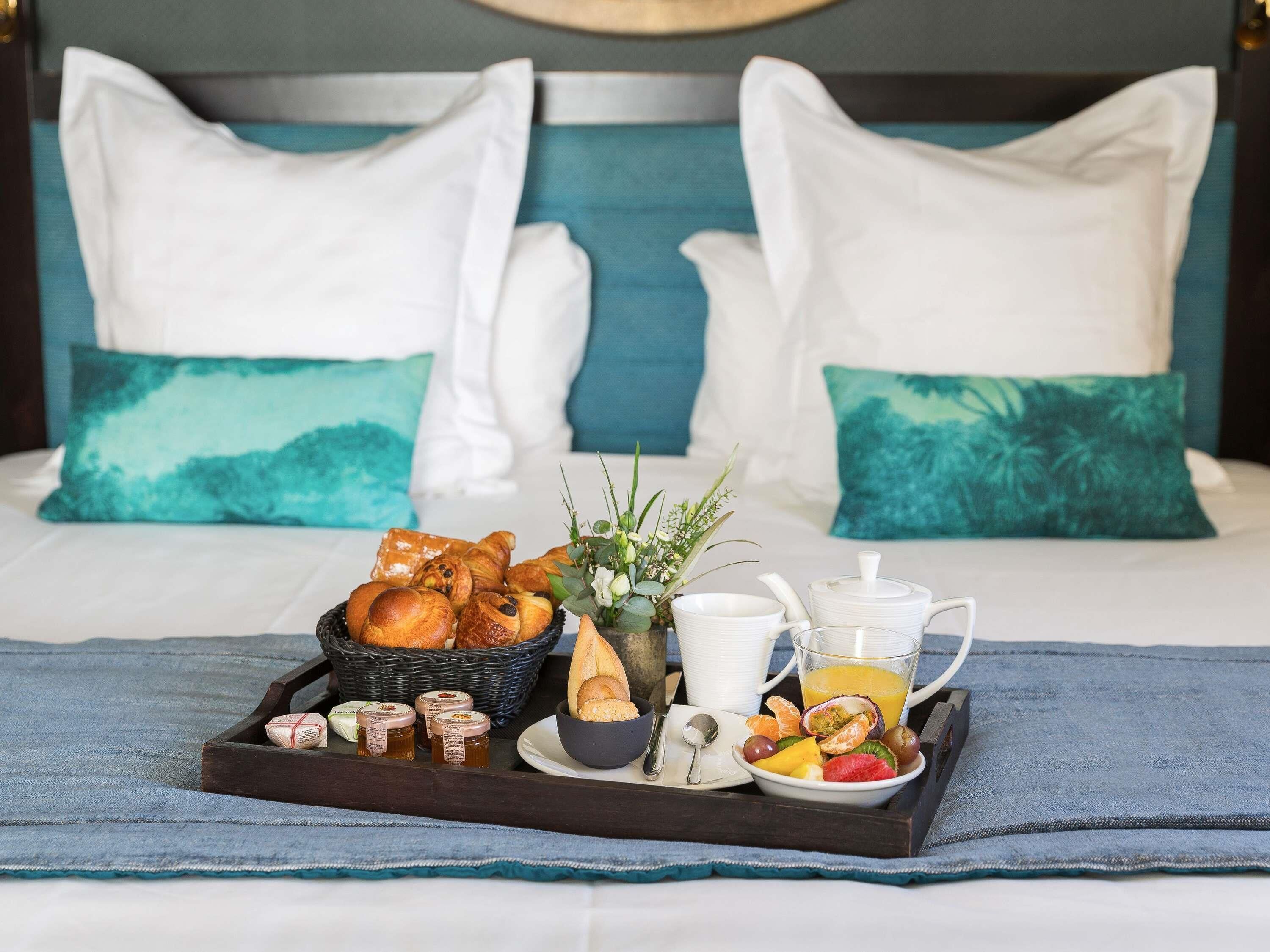 Bandeja de desayuno con pasteles, frutas y condimentos sobre una cama en un ambiente azul y blanco.