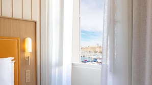 A hotel room with a bed, a nightstand, a wall-mounted light, and a window with a view of a marina and buildings. - Grand Hotel Beauvau Marseille Vieux-Port - MGallery Collection (Marseille)