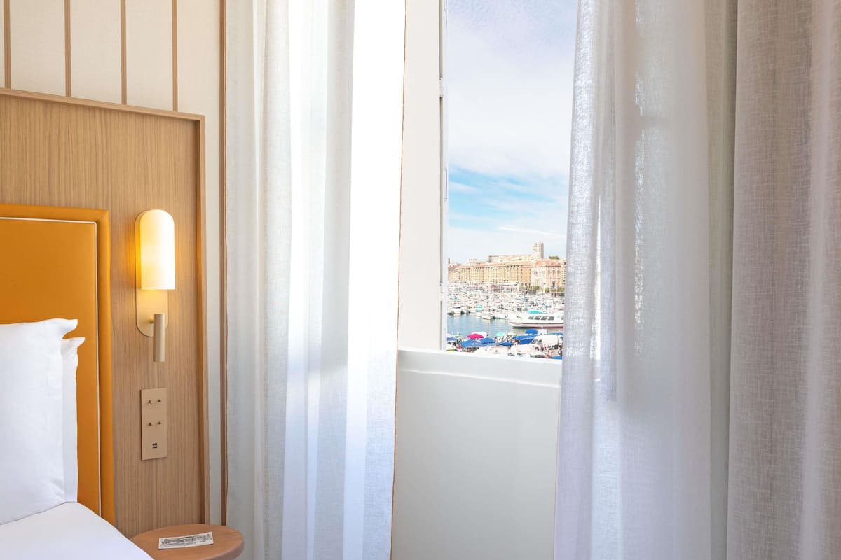 Et hotelværelse med en seng, et natbord, en vægmonteret lampe og et vindue med udsigt over en lysthavn og bygninger.