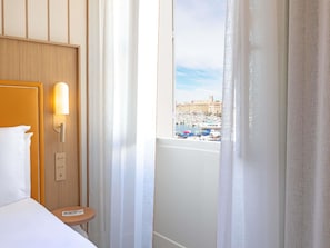 Et hotelværelse med en seng, et natbord, en vægmonteret lampe og et vindue med udsigt over en lysthavn og bygninger.