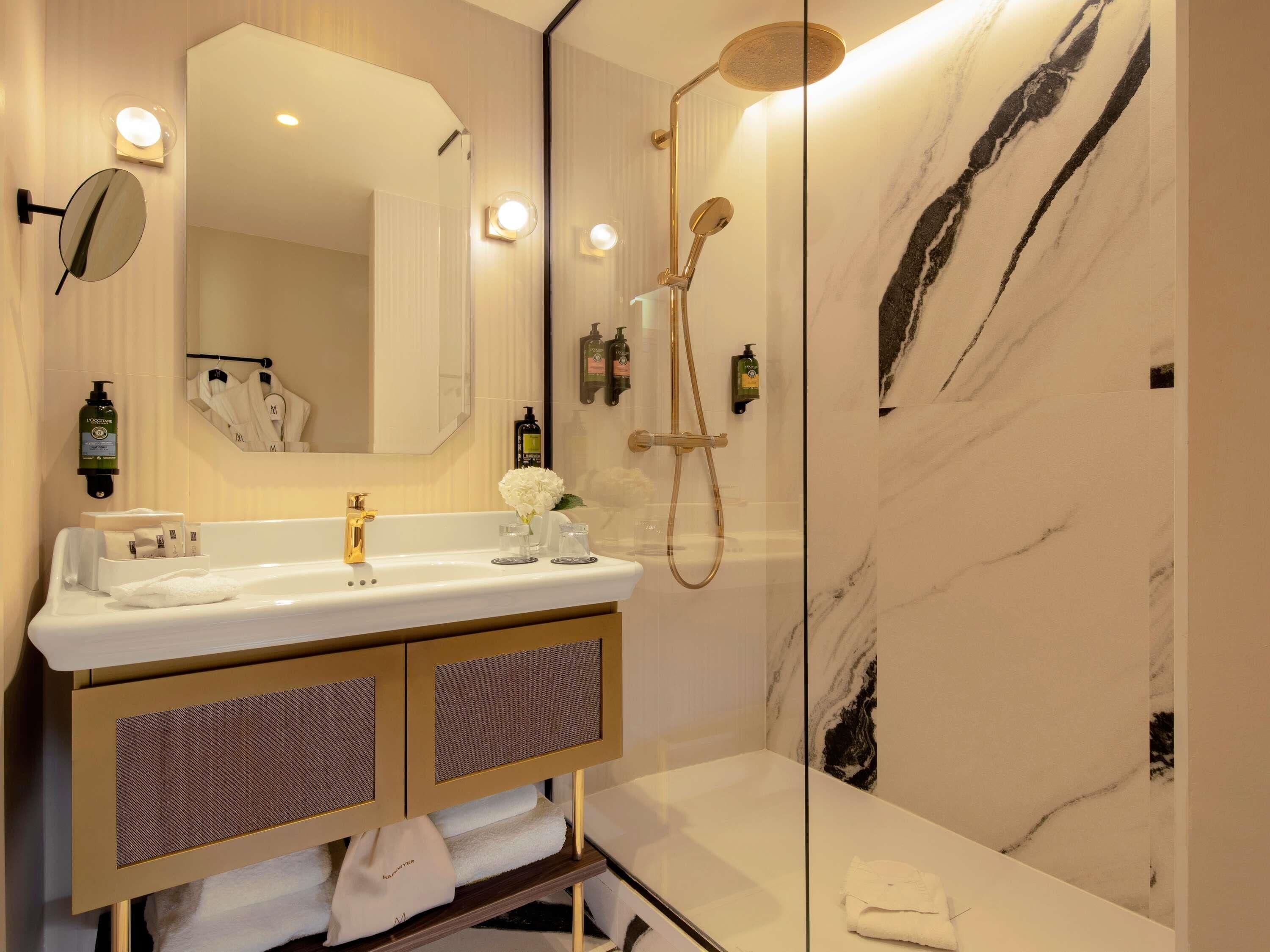 Un baño moderno con pared de mármol, tocador dorado, espejo grande y ducha con cabezal de ducha manual.