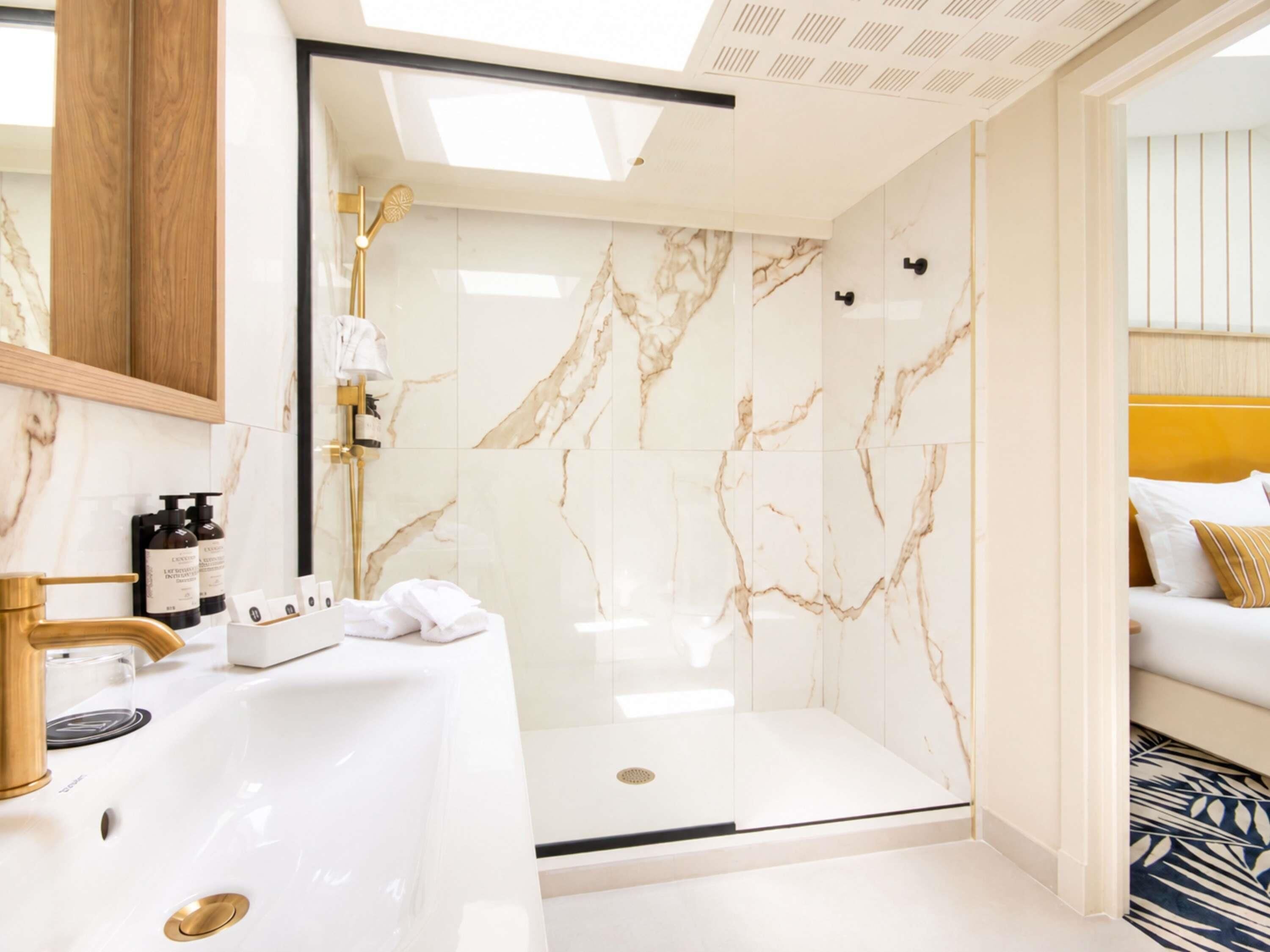 Un baño moderno con ducha de mármol blanco, grifo dorado y lavamanos blanco.