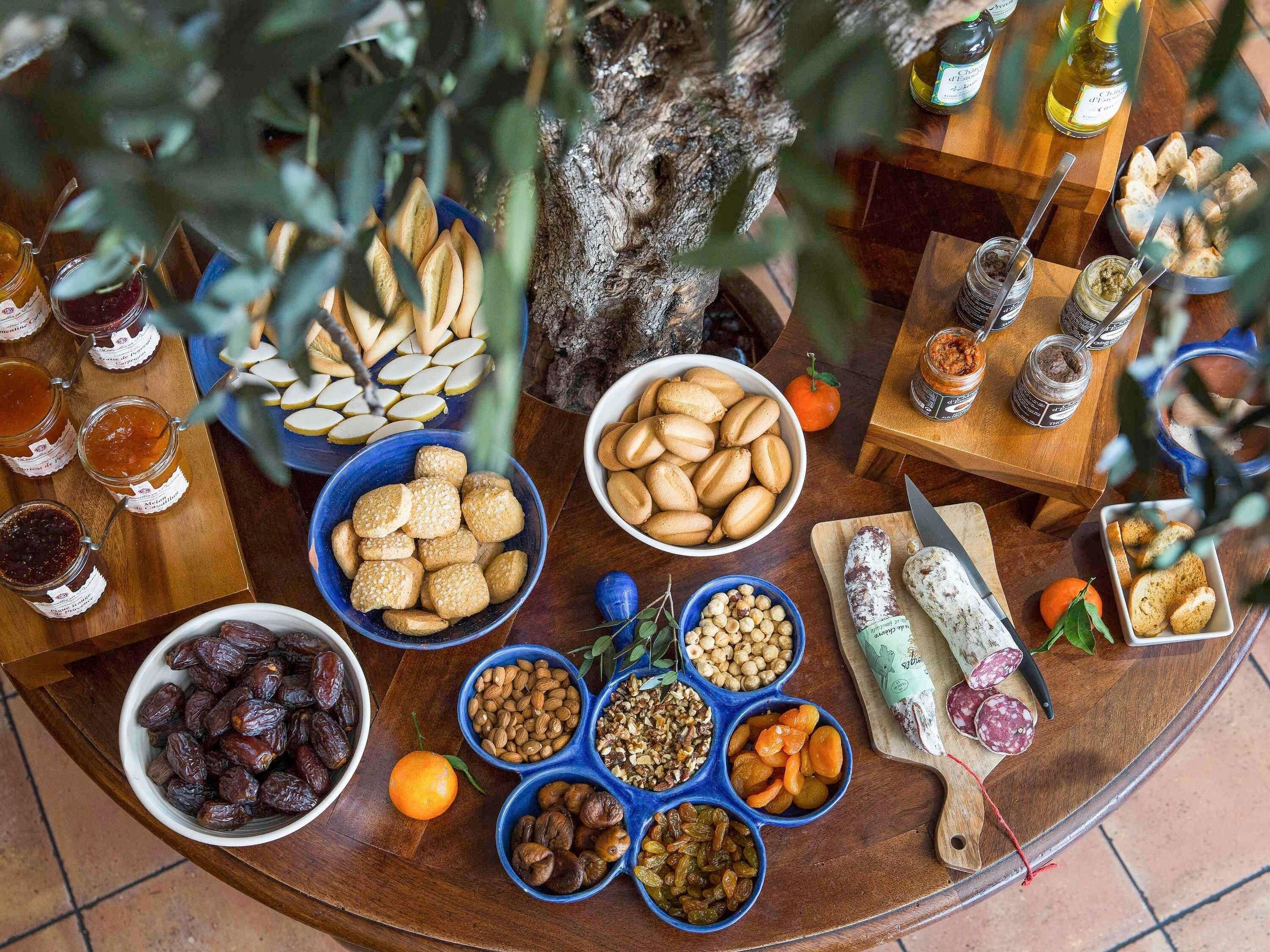A variety of food items and condiments arranged on a wooden table, including jars of preserves, nuts, and dried fruits.