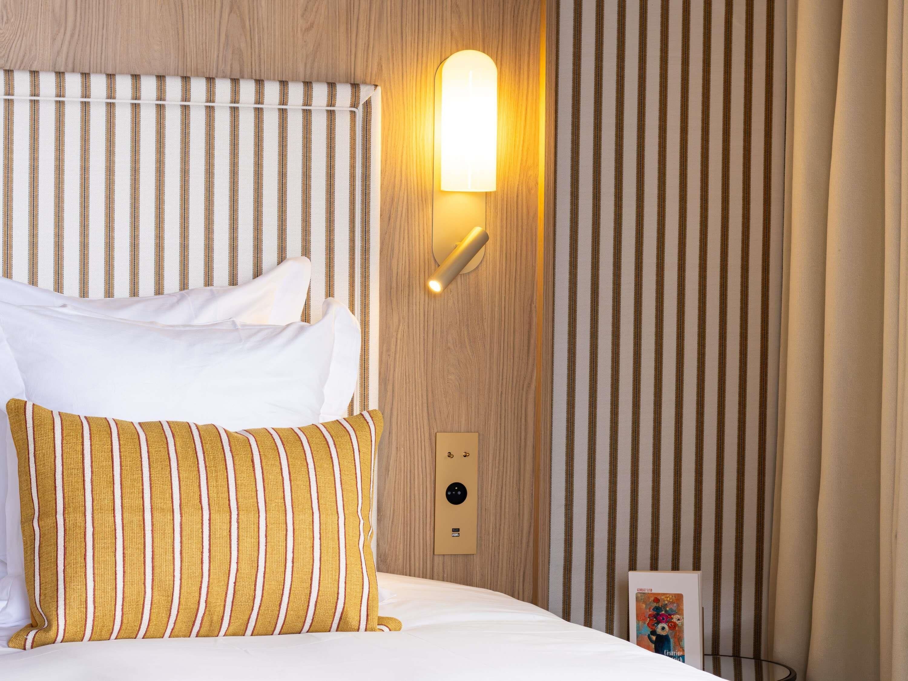 Een hotelkamer met een bed, gestreepte gordijnen, een wandlamp en een klein tafeltje met een boek.