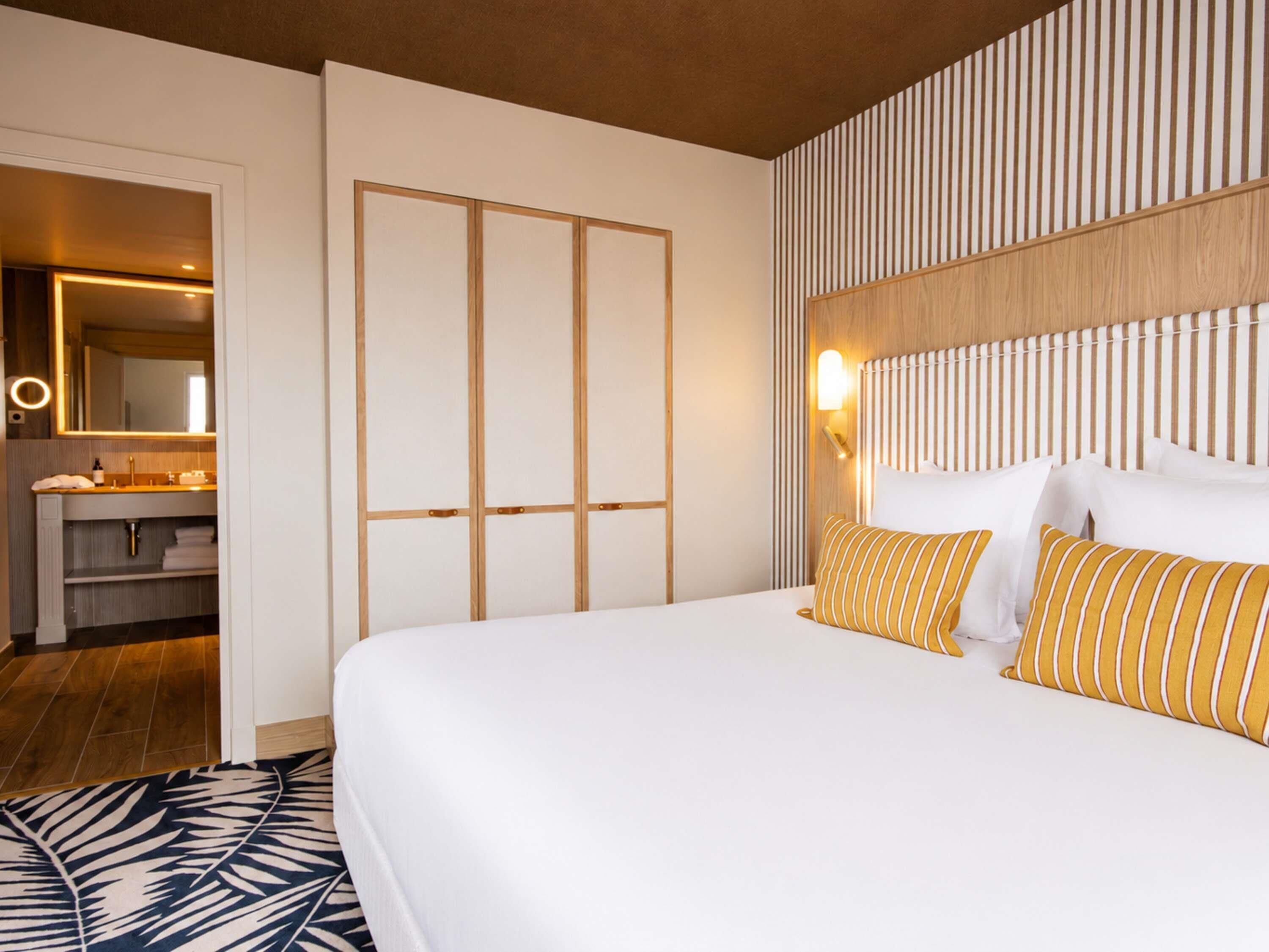 A hotel room with a bed, a bedside table, a mirror, and a bathroom visible in the background.