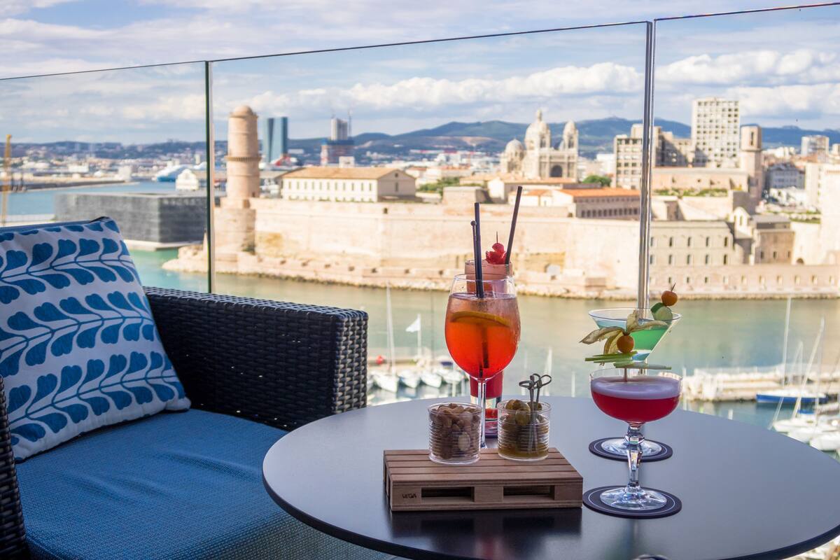 Un balcón con vistas a un paisaje urbano histórico, un sofá de mimbre con un cojín estampado con corazones, y dos bebidas sobre una mesita.