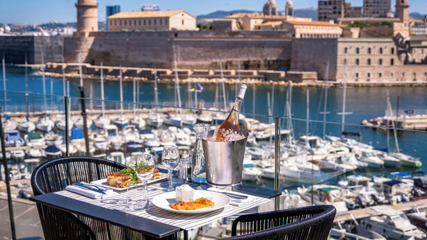 A table with a meal, a drink, and a view of a marina with boats and historic buildings. - Sofitel Marseille Vieux Port (Marseille)