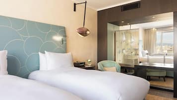 A hotel room with two beds, a desk, a chair, a large window with a view, and a wall-mounted lamp.