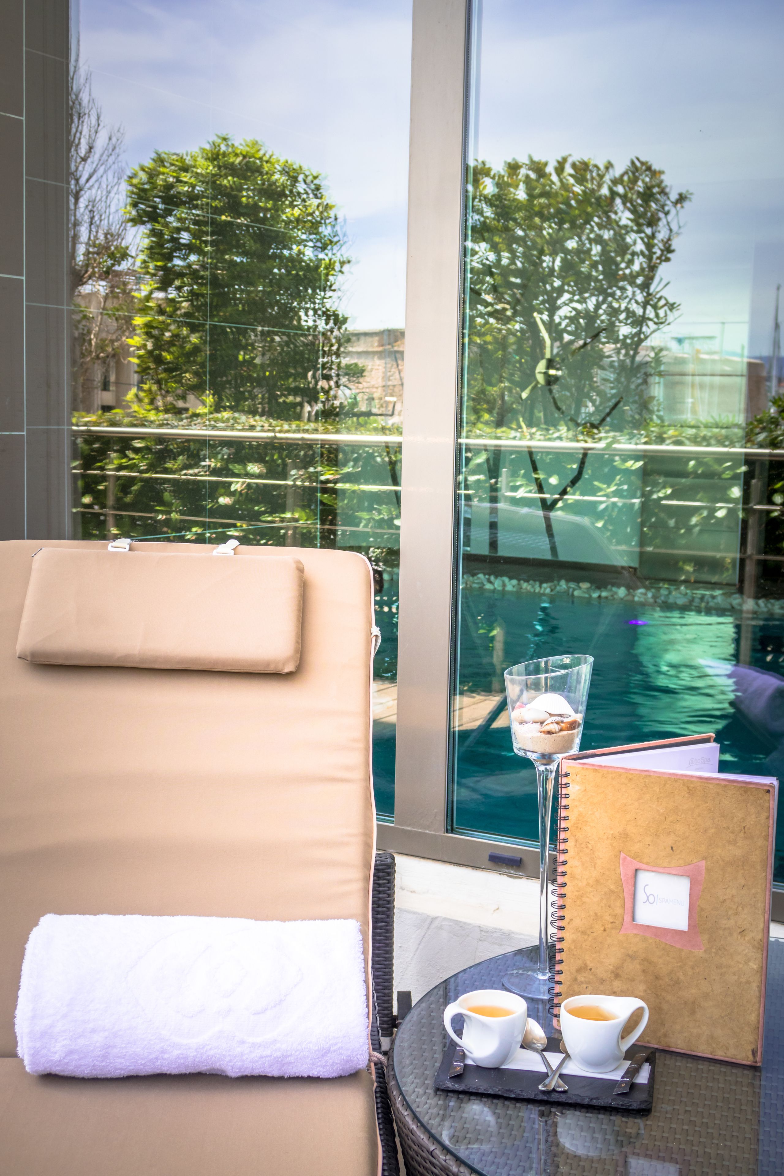 A sun lounger with a towel, a glass of iced coffee, and a notebook on a table by a window overlooking a pool.