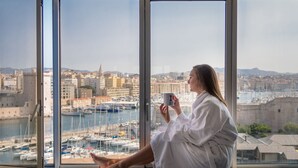A person sitting by a large window with a view of a marina and cityscape. - Sofitel Marseille Vieux Port (Marseille)