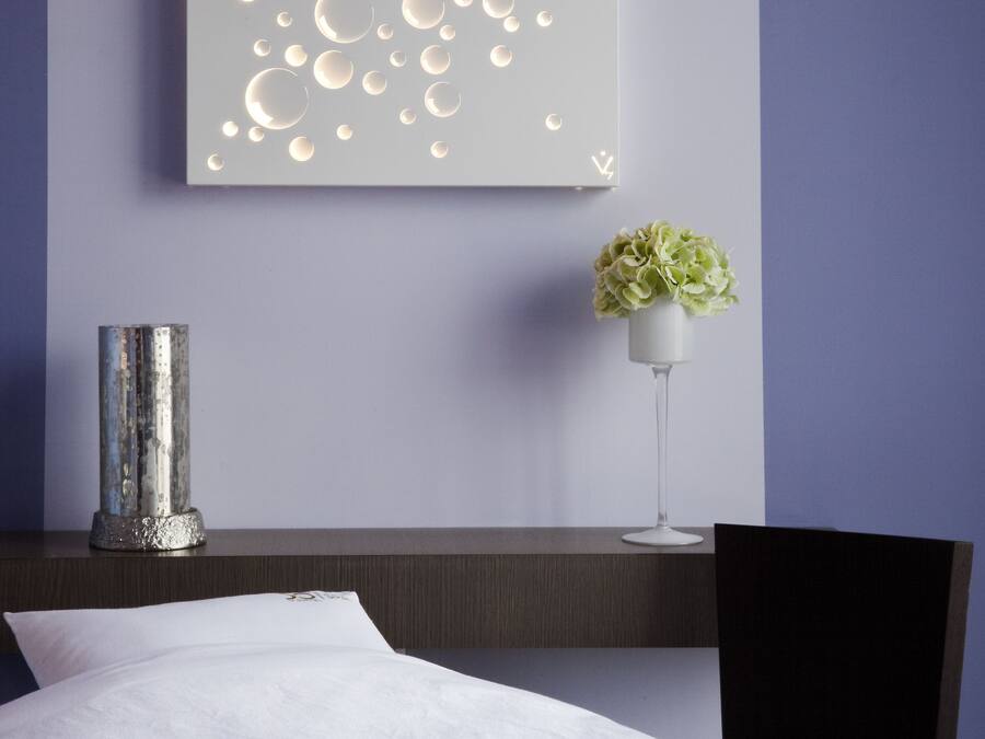 Una habitación de hotel moderna con una obra de arte grande en la pared, una mesita de noche con un jarrón de flores, una cama con cubrecama blanco y una silla.