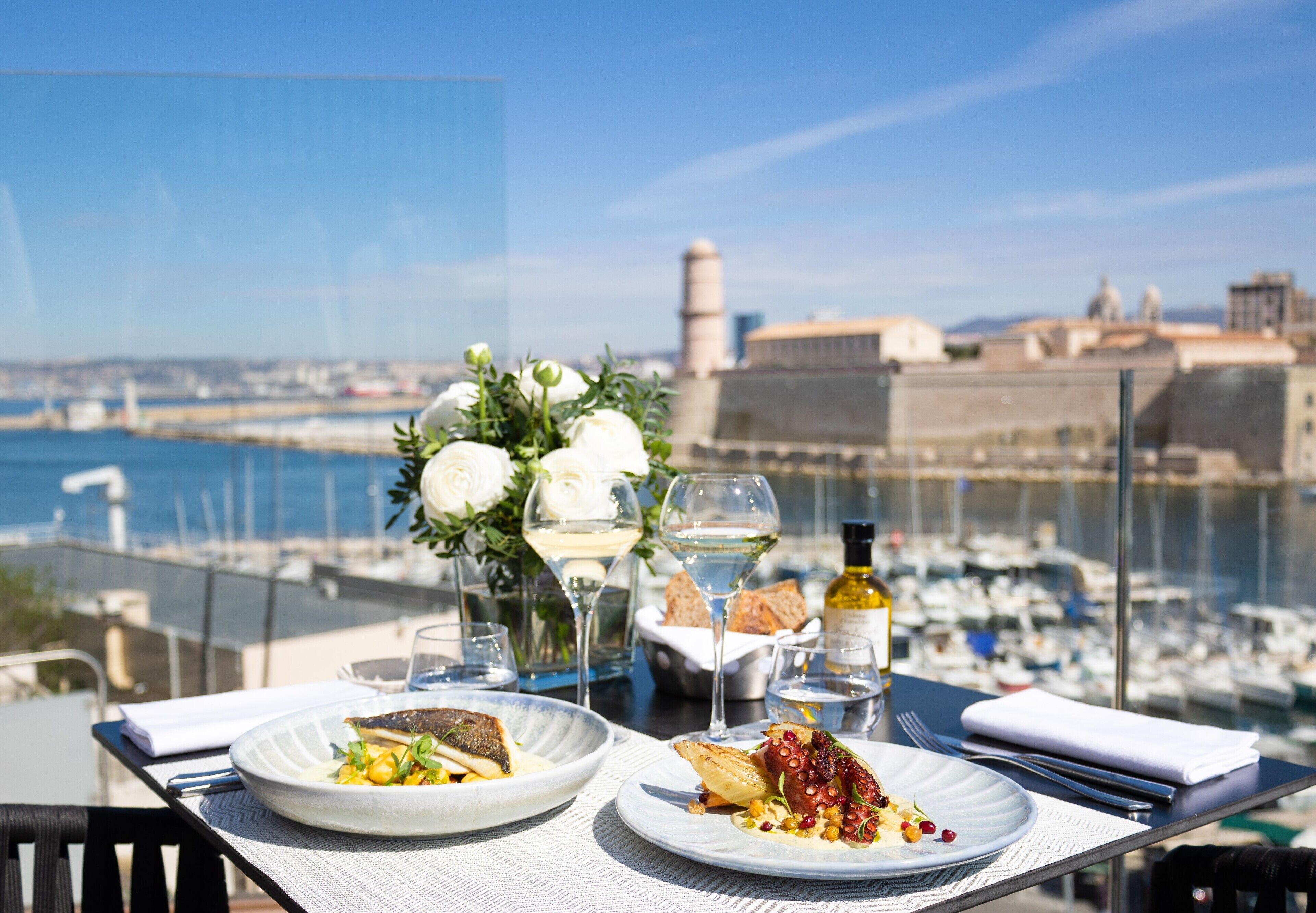 A dining table with plates of food, wine glasses, and a bottle of wine, overlooking a marina with boats and a cityscape in the background.