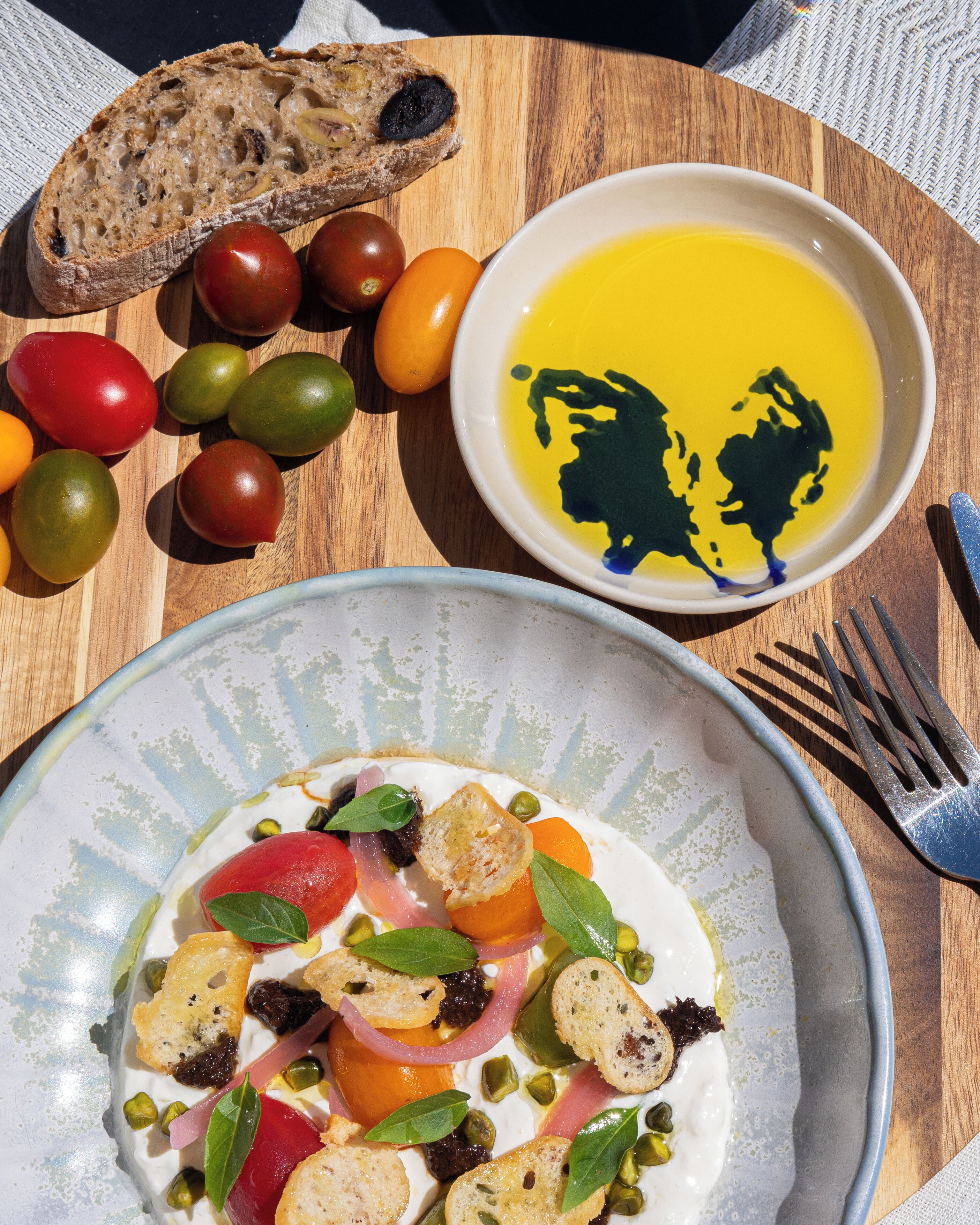 Una mesa de madera con un plato de comida, un tazón de aceite de oliva y una variedad de tomates de colores.