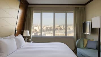 A hotel room with a large bed, a television, a sofa, and a view of a cityscape through a large window.