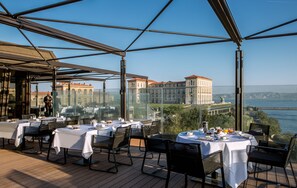 Un espace de restauration en plein air avec des tables disposées pour deux personnes, offrant une vue sur un plan d’eau et un bâtiment historique.