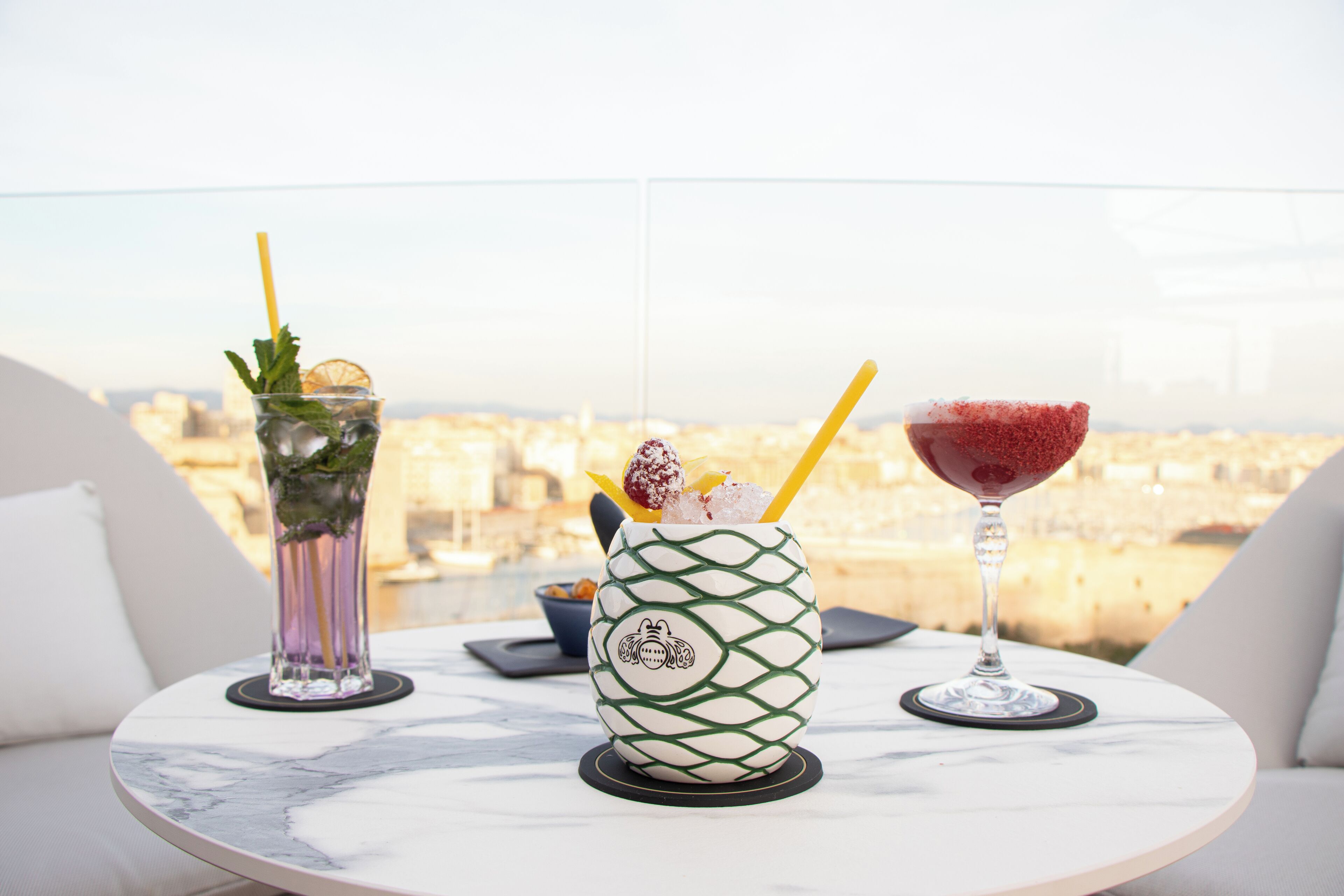 A table with three drinks on a balcony overlooking a cityscape.