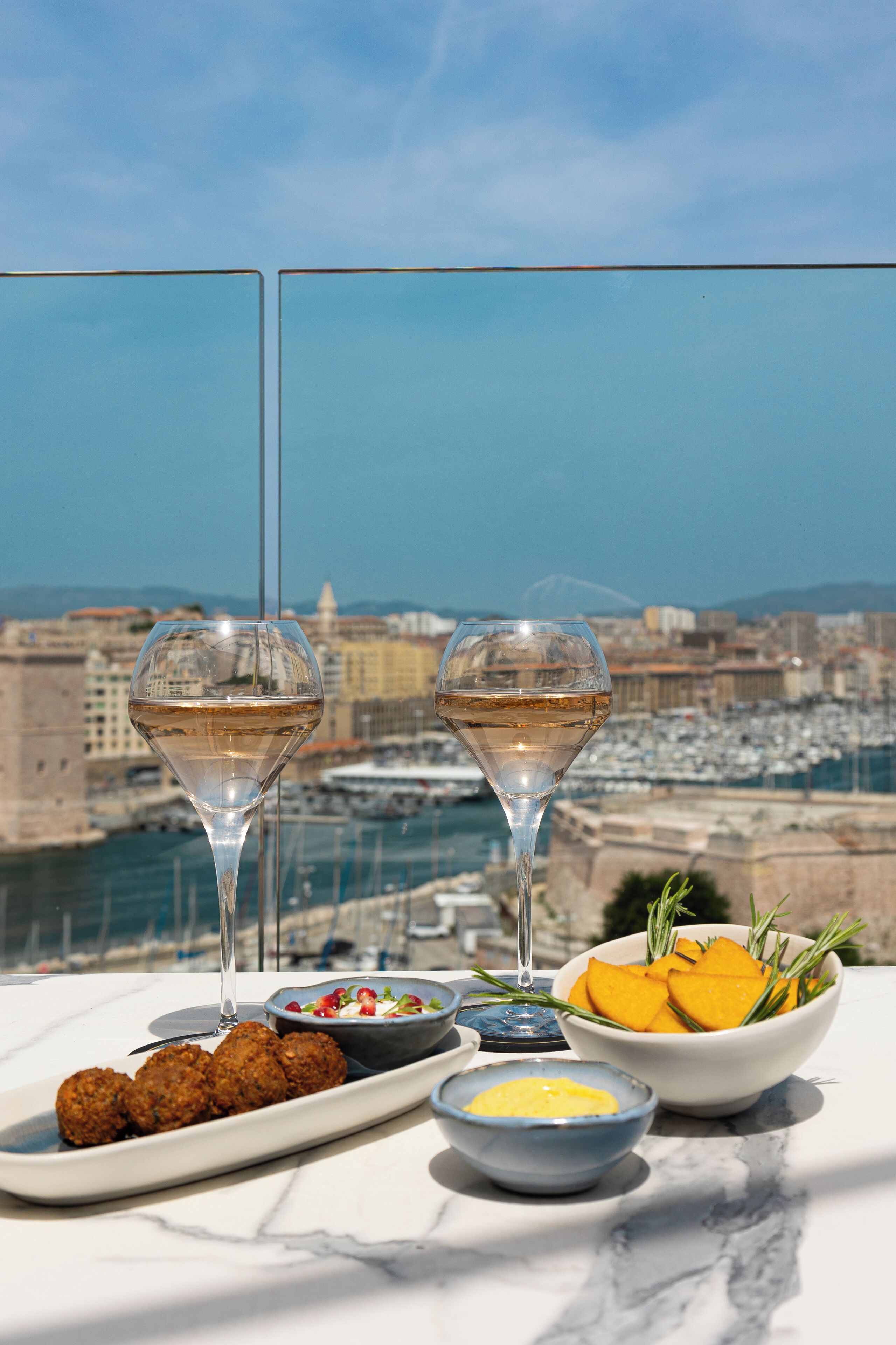 A table with a view of a marina and cityscape, featuring a plate of falafel, a bowl of salad, a bowl of fruit, and two glasses of white wine.