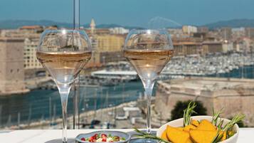 A table with a view of a marina and cityscape, featuring a plate of falafel, a bowl of salad, a bowl of fruit, and two glasses of white wine.