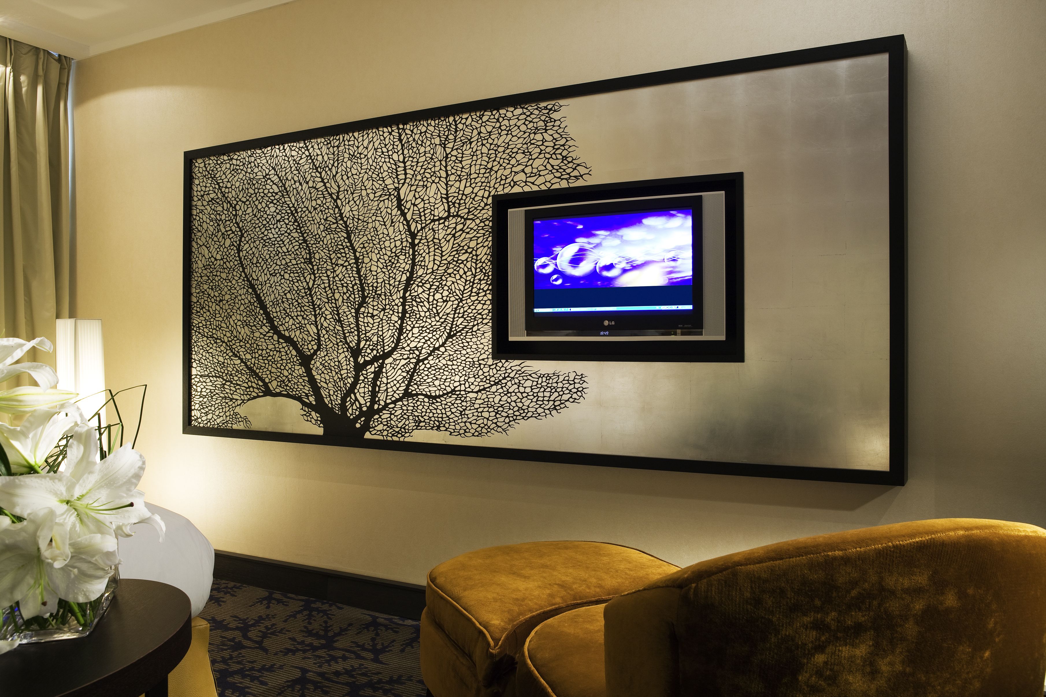 Habitación de hotel moderna con un gran cuadro de un árbol, un televisor de pantalla plana, un sillón amarillo mullido y un jarrón con flores blancas.