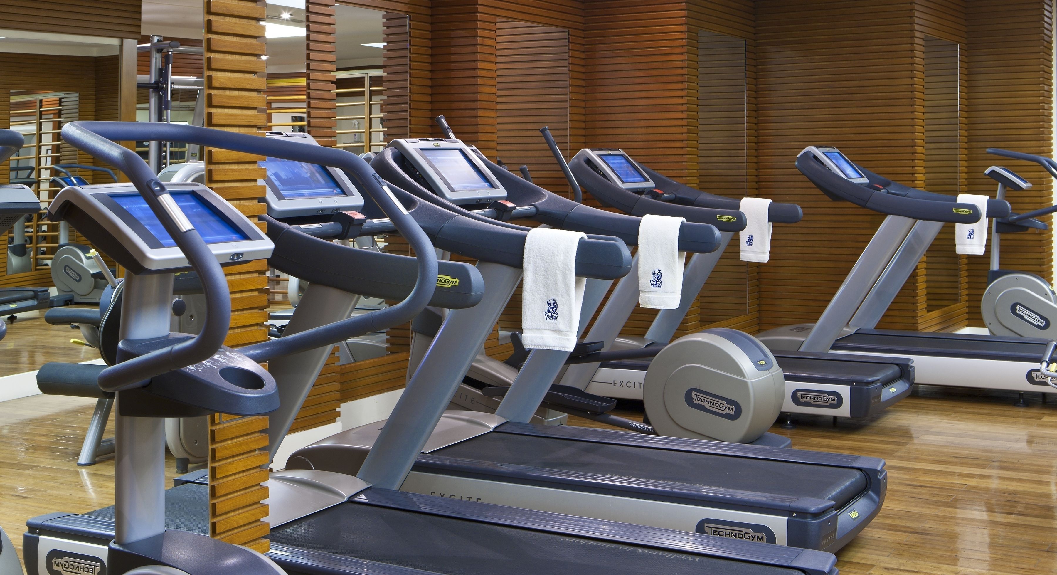 A row of Technogym treadmills in a gym with wooden paneling and a mirrored wall.