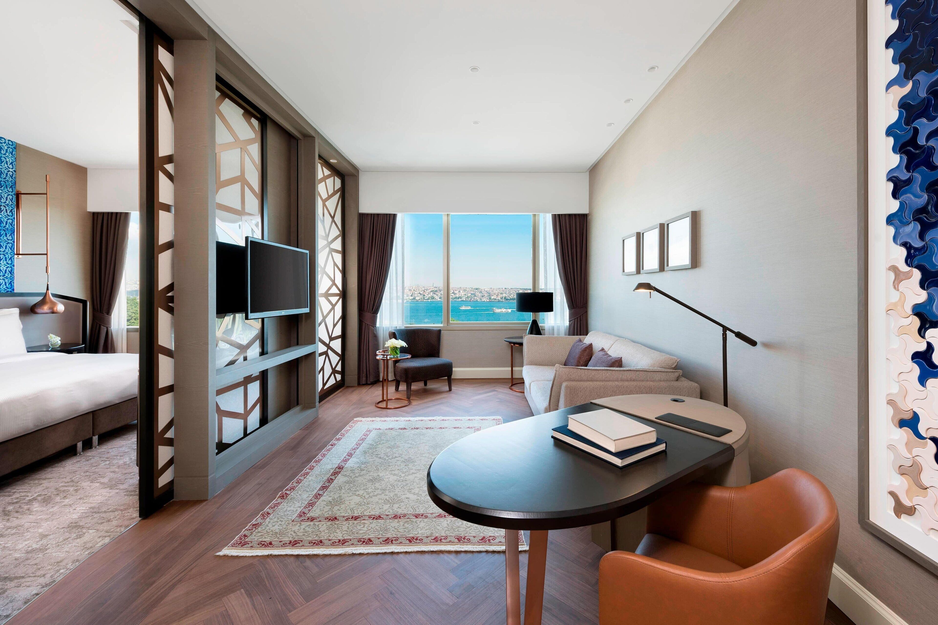 Quarto de hotel moderno com uma janela grande, um sofá, uma mesa de jantar e uma televisão.