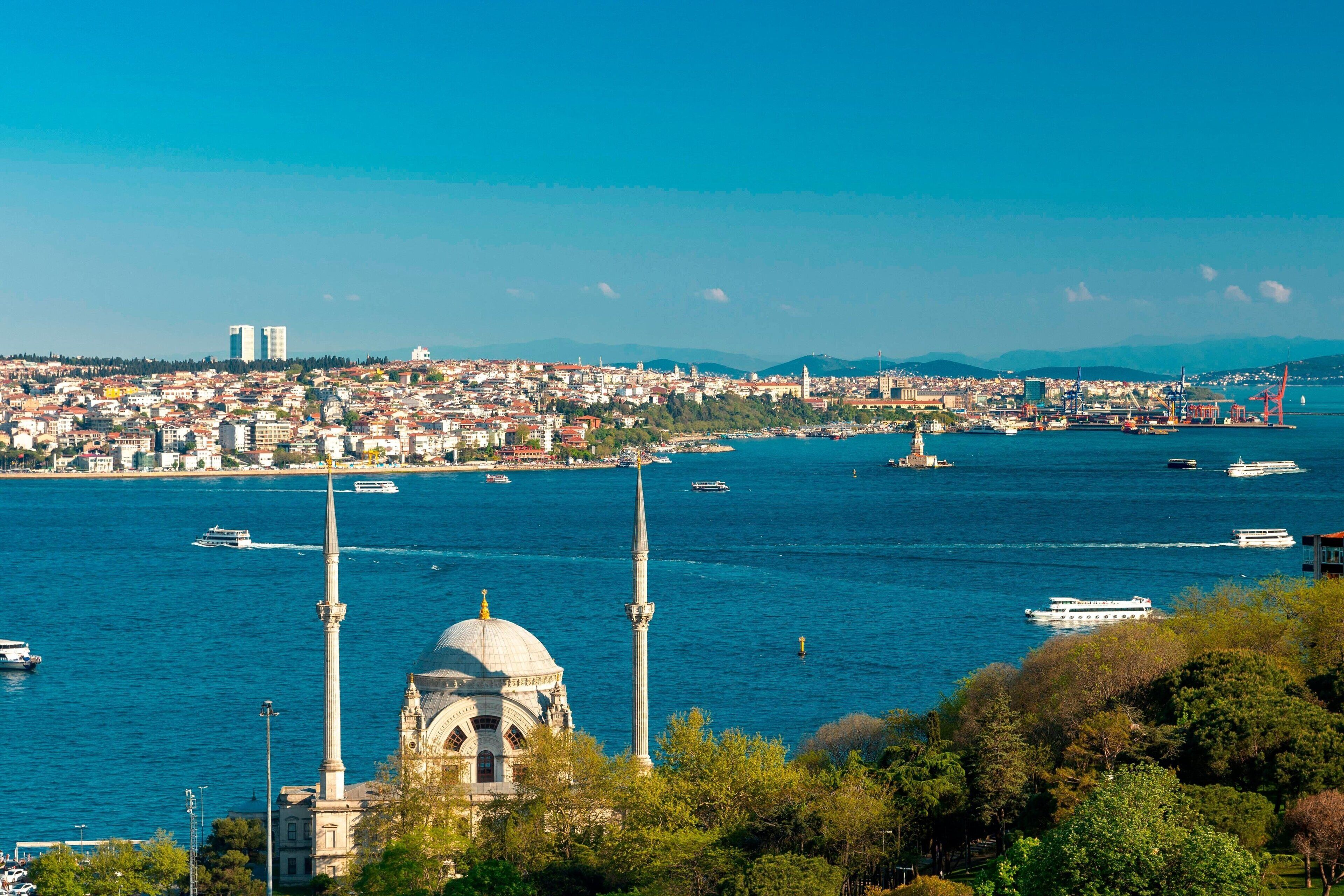A coastal cityscape with a prominent mosque featuring two minarets and a dome, surrounded by water and boats.