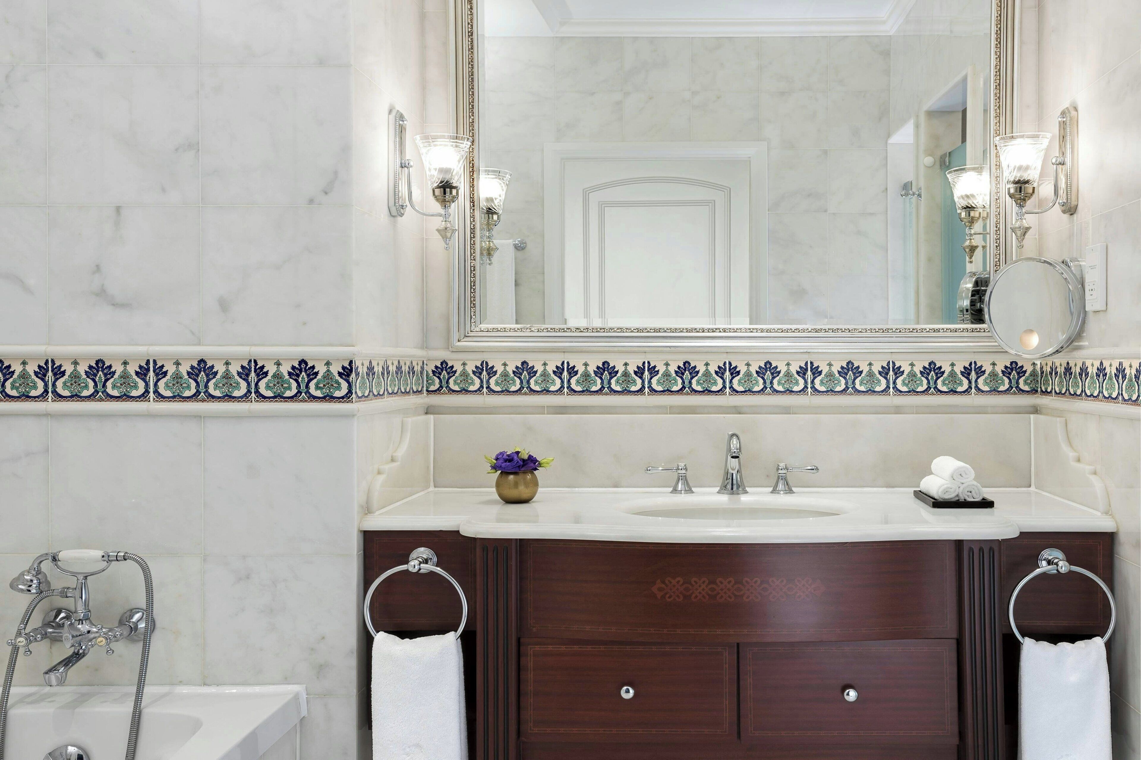 A bathroom with a marble wall, a large mirror, a sink, a bathtub, and two towel rings.