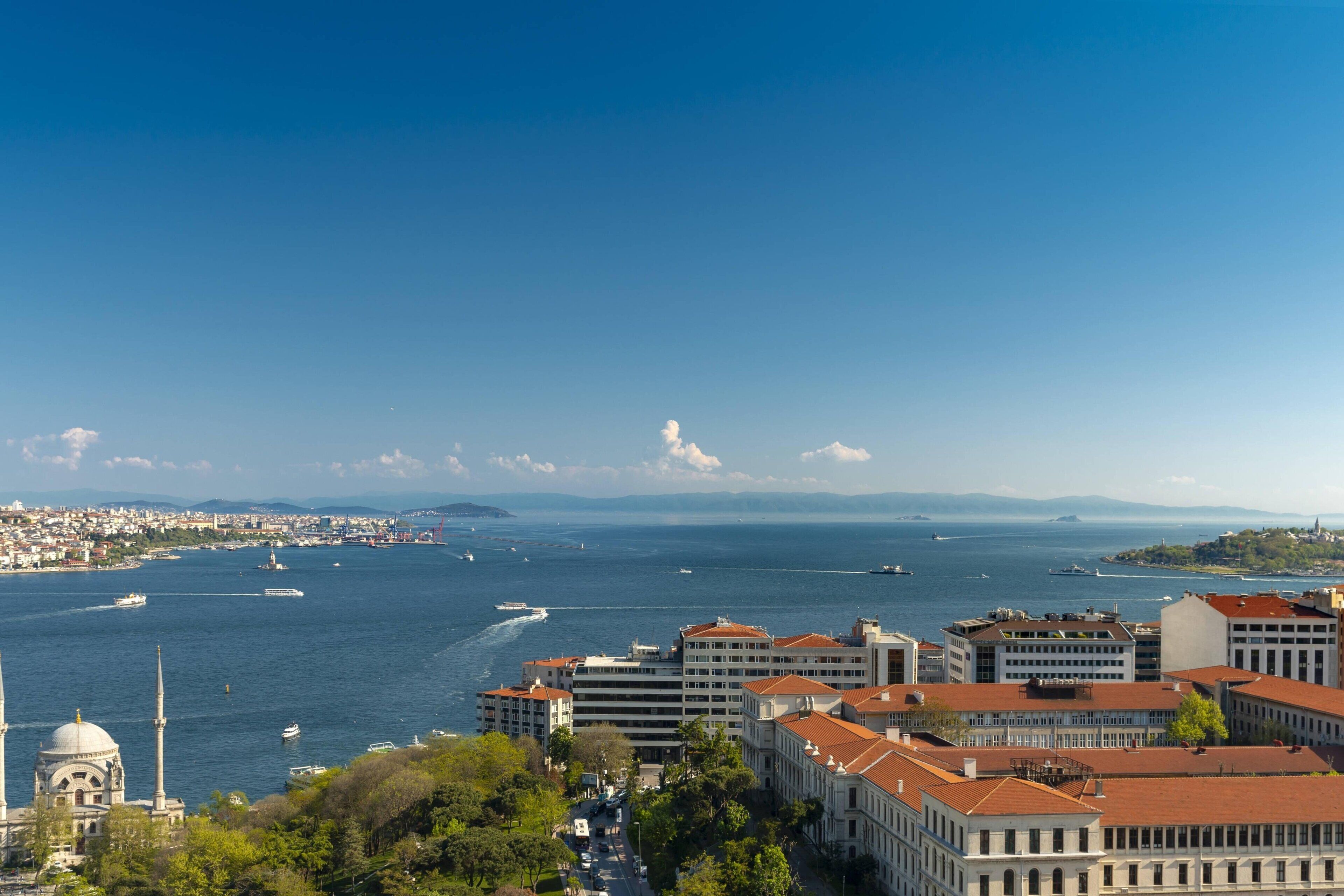 A coastal cityscape with a prominent mosque, buildings with red-tiled roofs, and a view of the sea with boats.