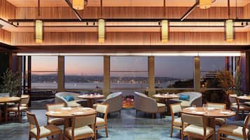Un restaurante con mesas y sillas de madera, amplios ventanales que ofrecen vistas de un paisaje urbano al anochecer.
