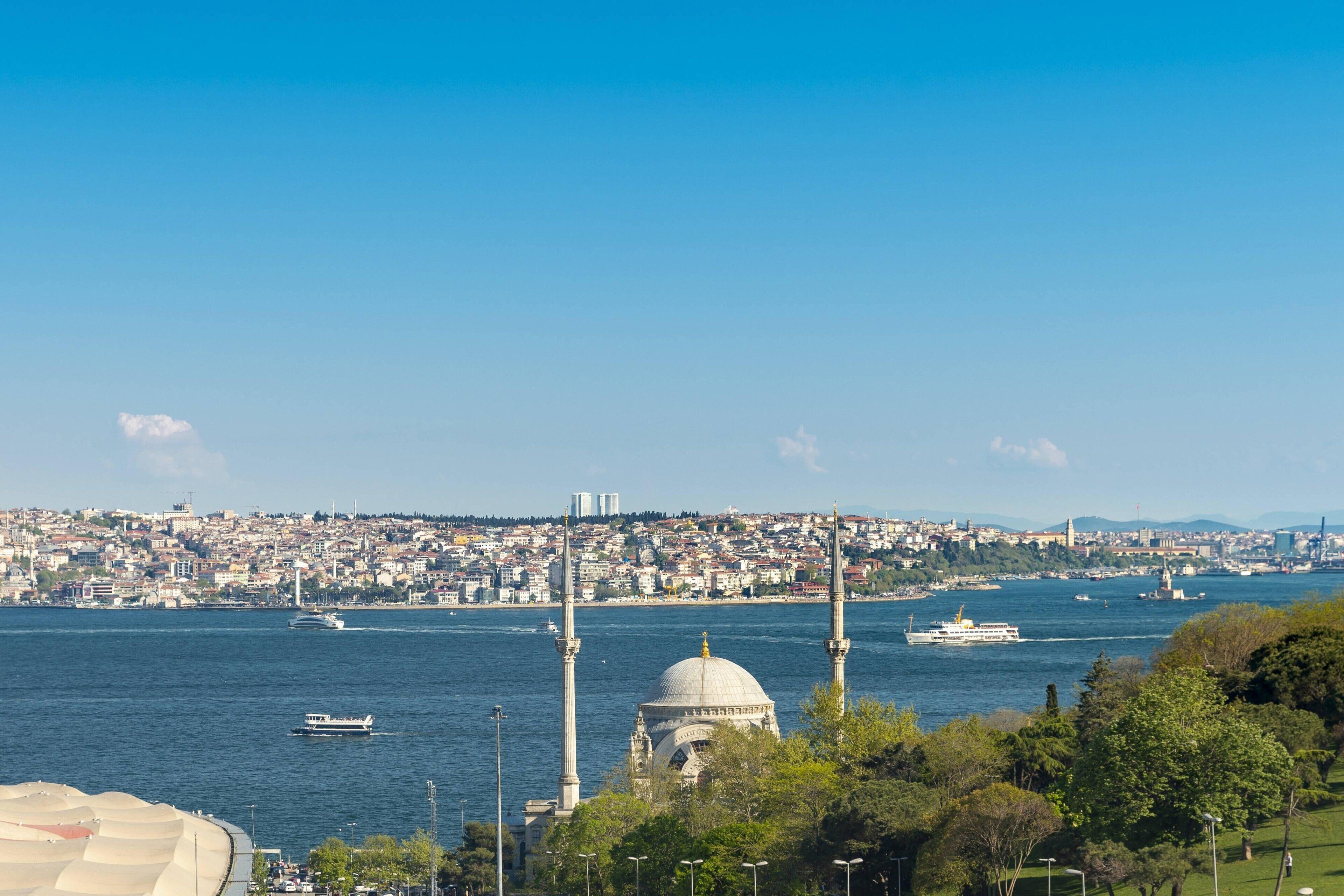 A cityscape with a prominent mosque with a dome and minarets, a body of water with boats, and a densely populated urban area in the background.