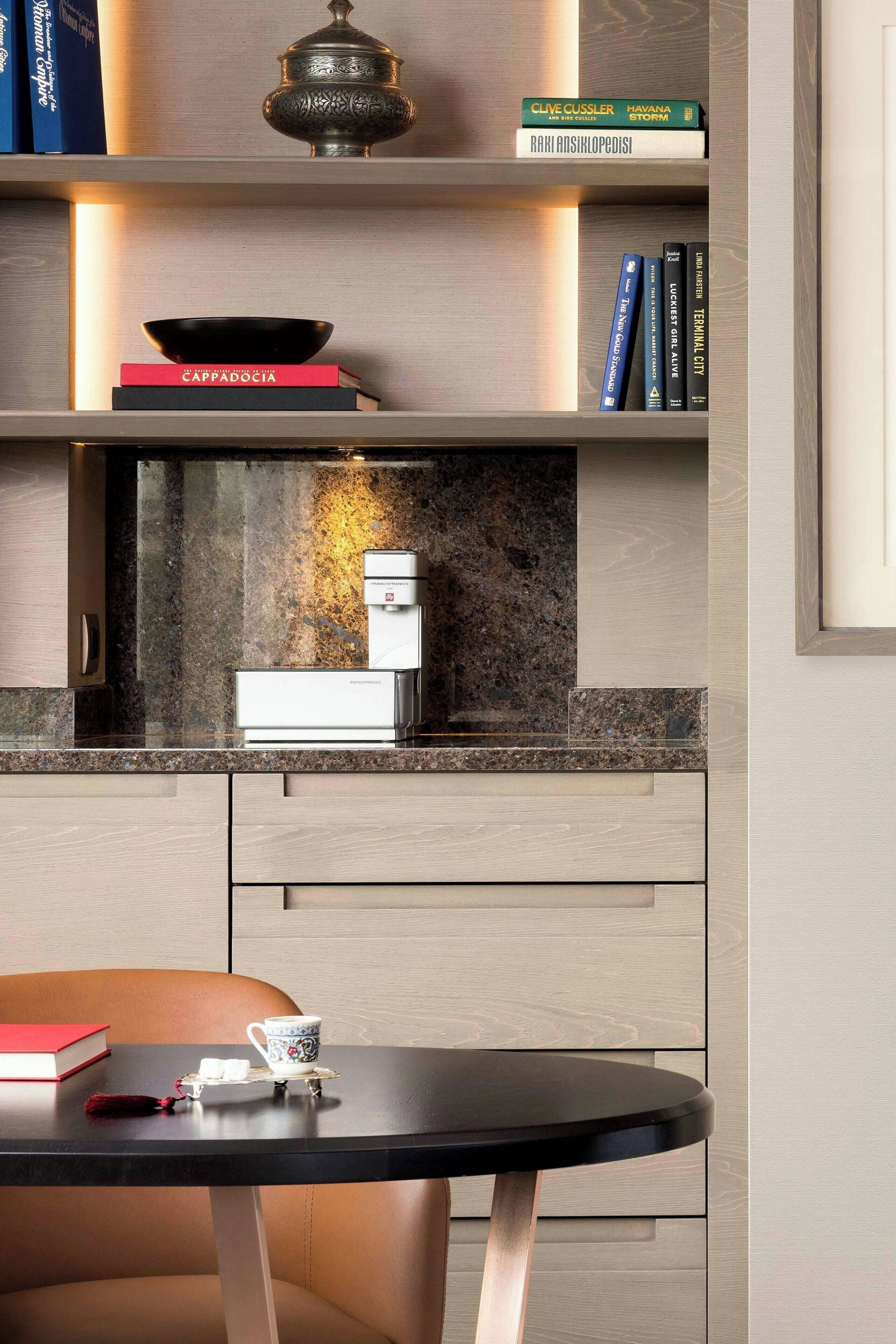 A modern kitchen with a built-in coffee machine, a black table with a teapot and cups, and a bookshelf with books and decorative items.