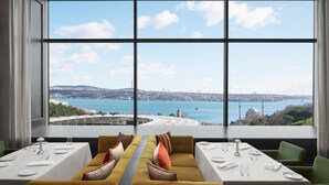 A dining area with a table set for two, a sofa with cushions, and a large window offering a view of a body of water and distant buildings.