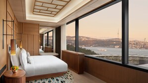 A modern hotel room with a large window, a bed, a bedside table, and a view of a cityscape and water.