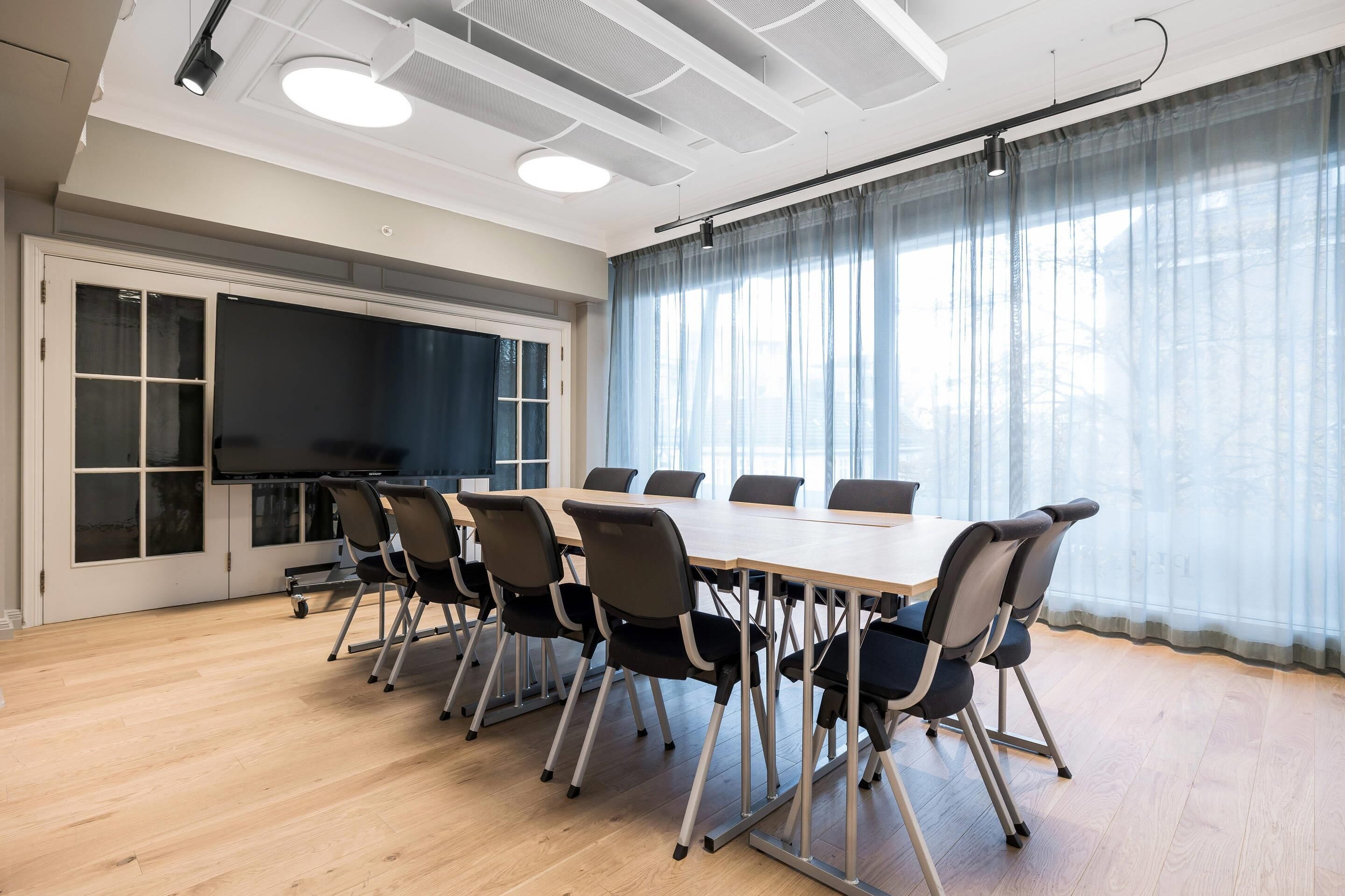 A conference room with a long table, chairs, a large screen, and a view through sheer curtains.