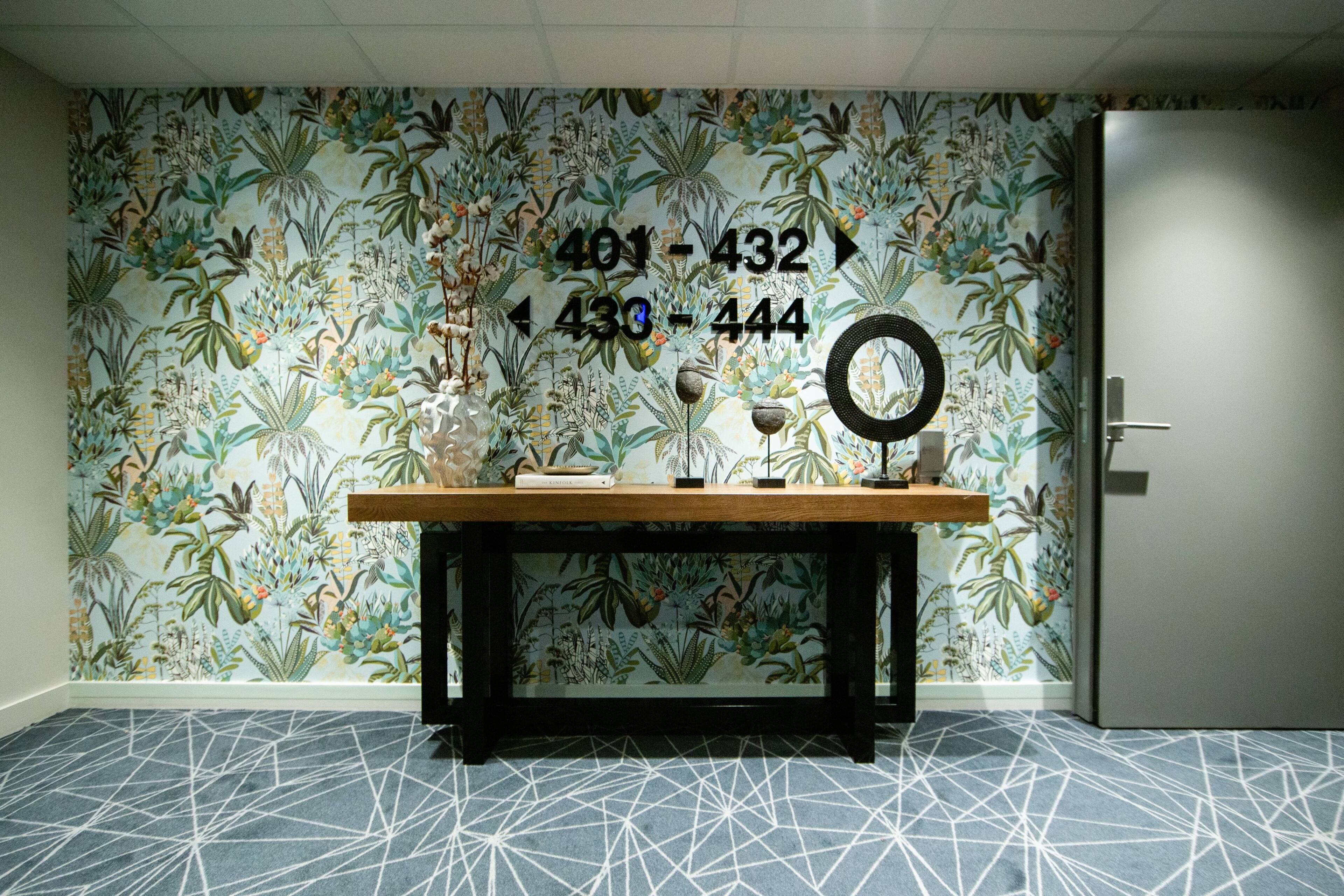 A room with a floral wallpaper, a wooden table with books and a mirror, and a geometric patterned carpet.