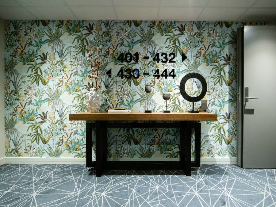 A room with a floral wallpaper, a wooden table with books and a mirror, and a geometric patterned carpet.