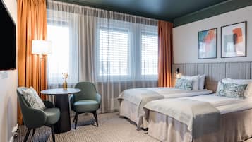 Habitación de hotel con dos camas, una mesa redonda con sillas, una lámpara y vistas a la ciudad a través de amplios ventanales.
