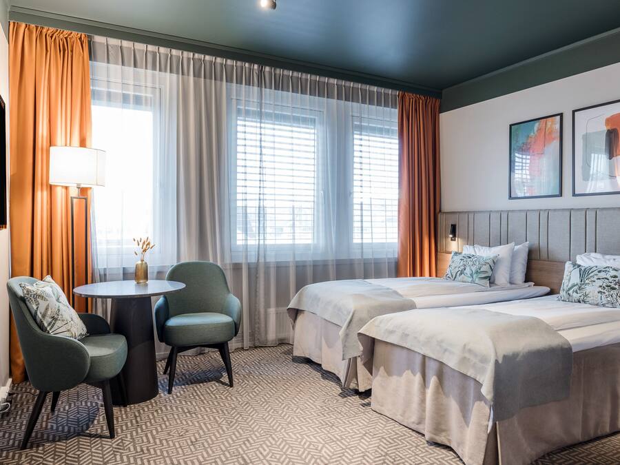 Habitación de hotel con dos camas, una mesa redonda con sillas, una lámpara y vistas a la ciudad a través de amplios ventanales.