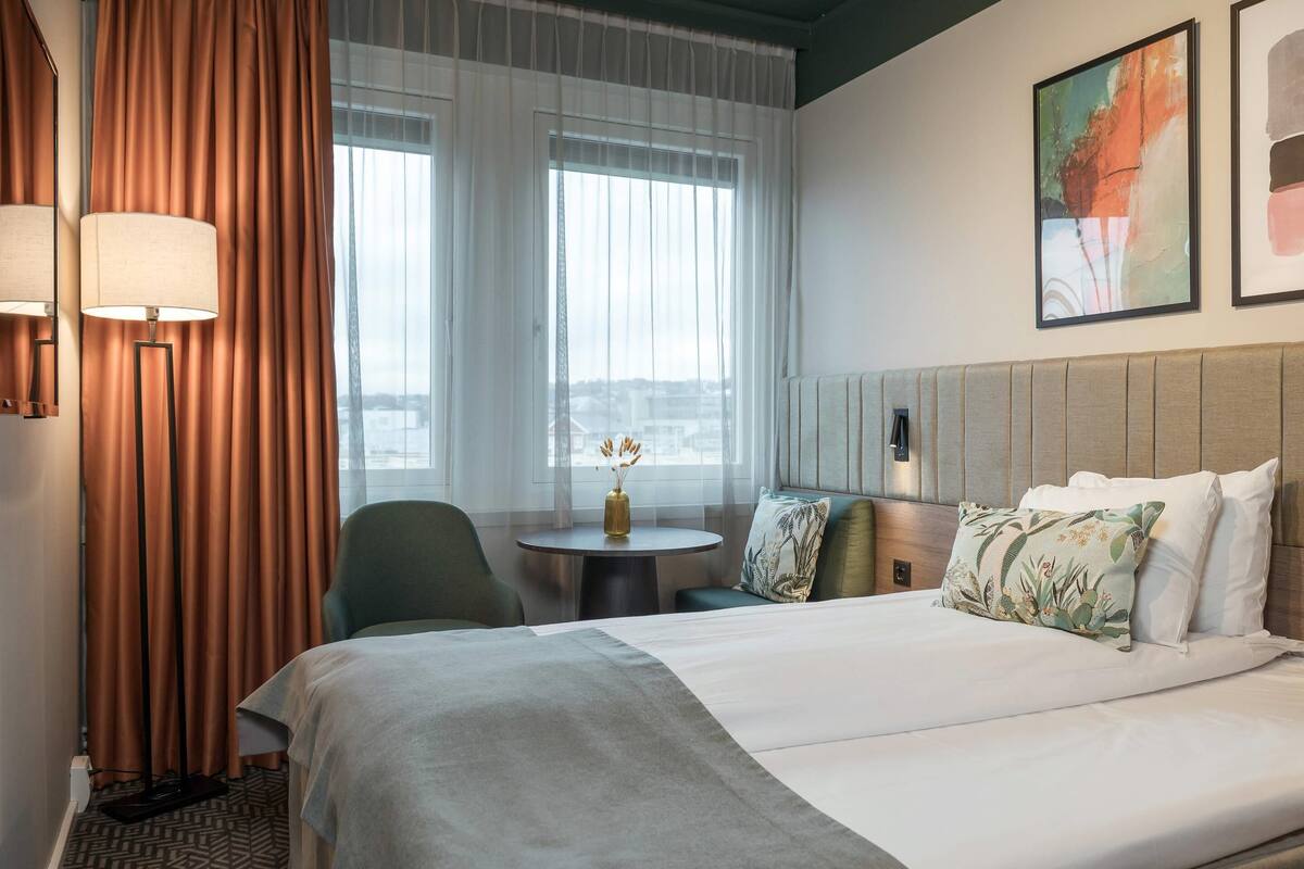 A hotel room with a bed, a round table with a vase, a chair, and a view of the city through large windows.