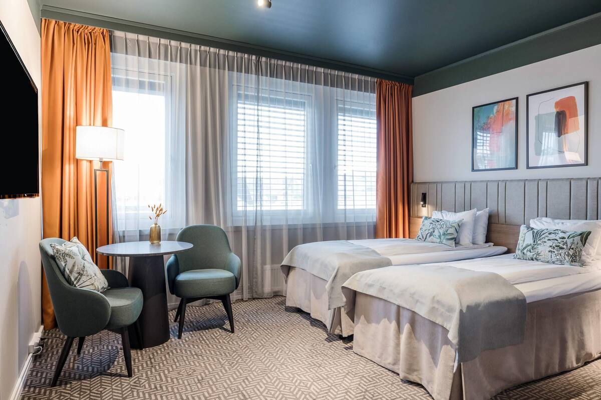 A hotel room with two beds, a round table, two chairs, a lamp, and large windows with curtains.