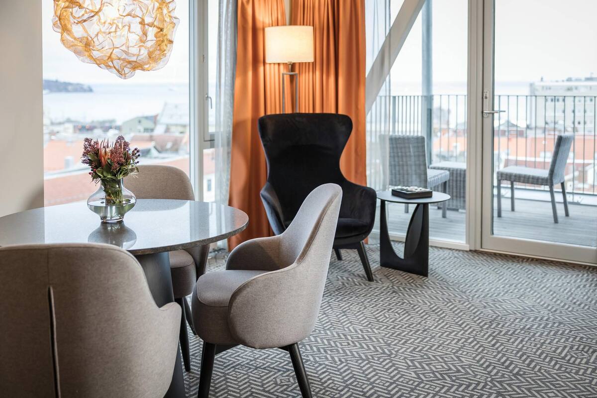 A modern hotel room with a round table, two chairs, a vase with flowers, a lamp, and a view of the city through large windows.