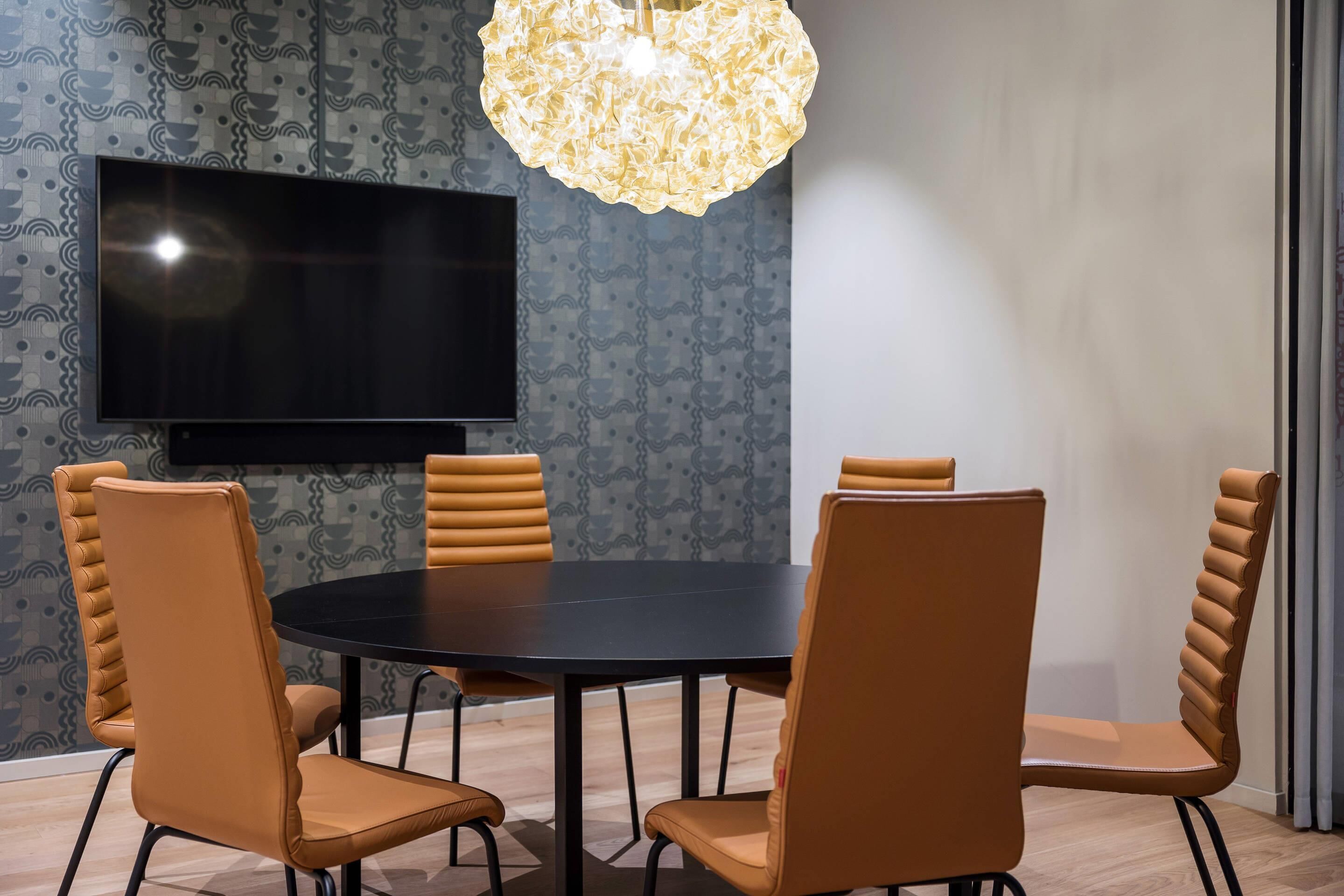 A conference room with a round table, four chairs, a flat-screen TV, and a patterned wallpaper.