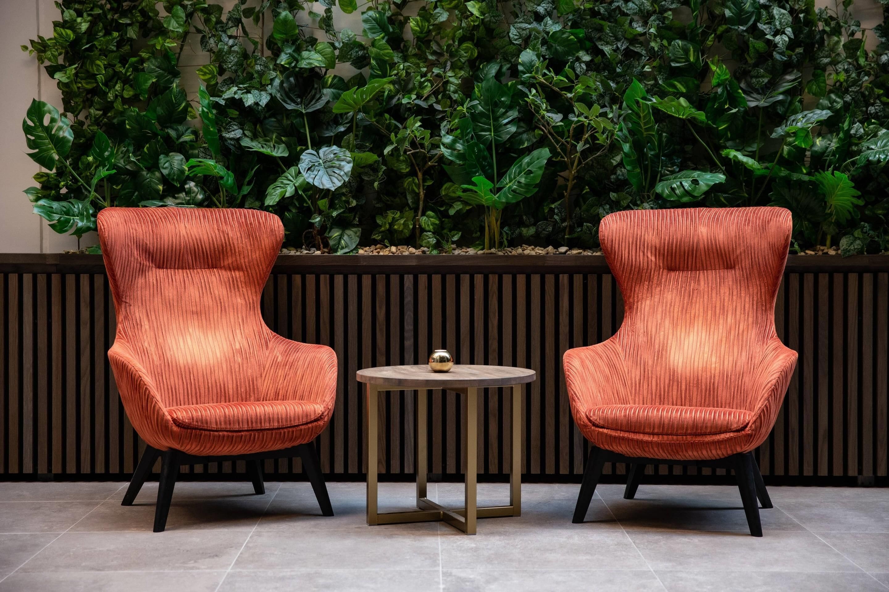 Dos sillones tapizados en naranja con patas negras, una mesita redonda con base dorada y una pared cubierta de follaje verde.
