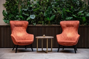Two orange upholstered chairs with black legs, a small round table with a gold base, and a wall of green foliage.