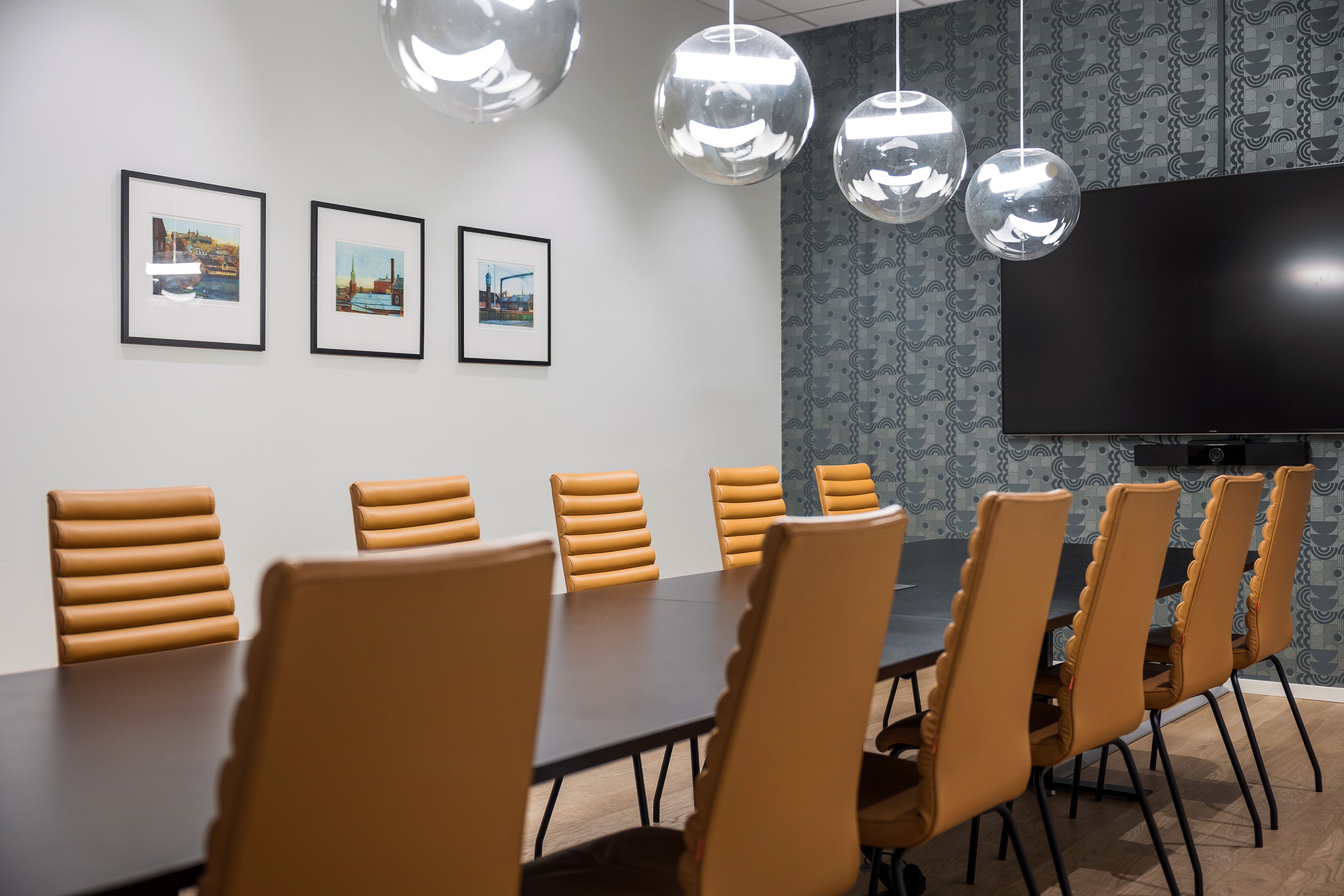 A conference room with a long table, multiple chairs, framed pictures on the wall, a large flat-screen TV, and hanging spherical light fixtures.