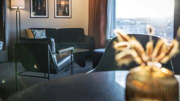 Habitación de hotel moderna con sofá, una mesita y un jarrón en primer plano. Se ven dos cuadros en la pared y un ventanal con vistas a la ciudad.
