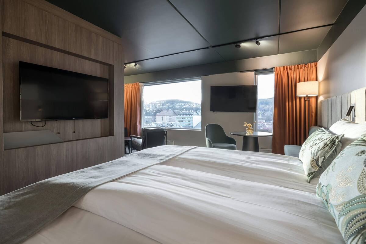 A hotel room with a large bed, a flat-screen TV mounted on the wall, a desk with a lamp, and a view of a cityscape through the window.