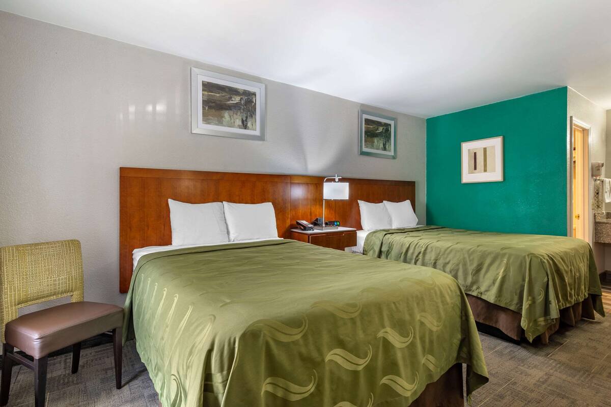 A hotel room with two beds, a wooden headboard, a green bedspread, a chair, and a nightstand with a lamp.