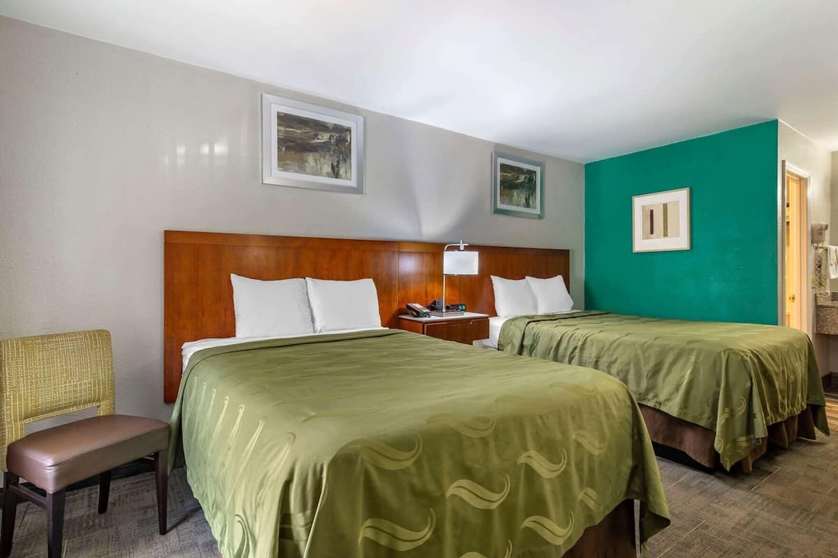A hotel room with two beds, a wooden headboard, a bench, a chair, and artwork on the walls.