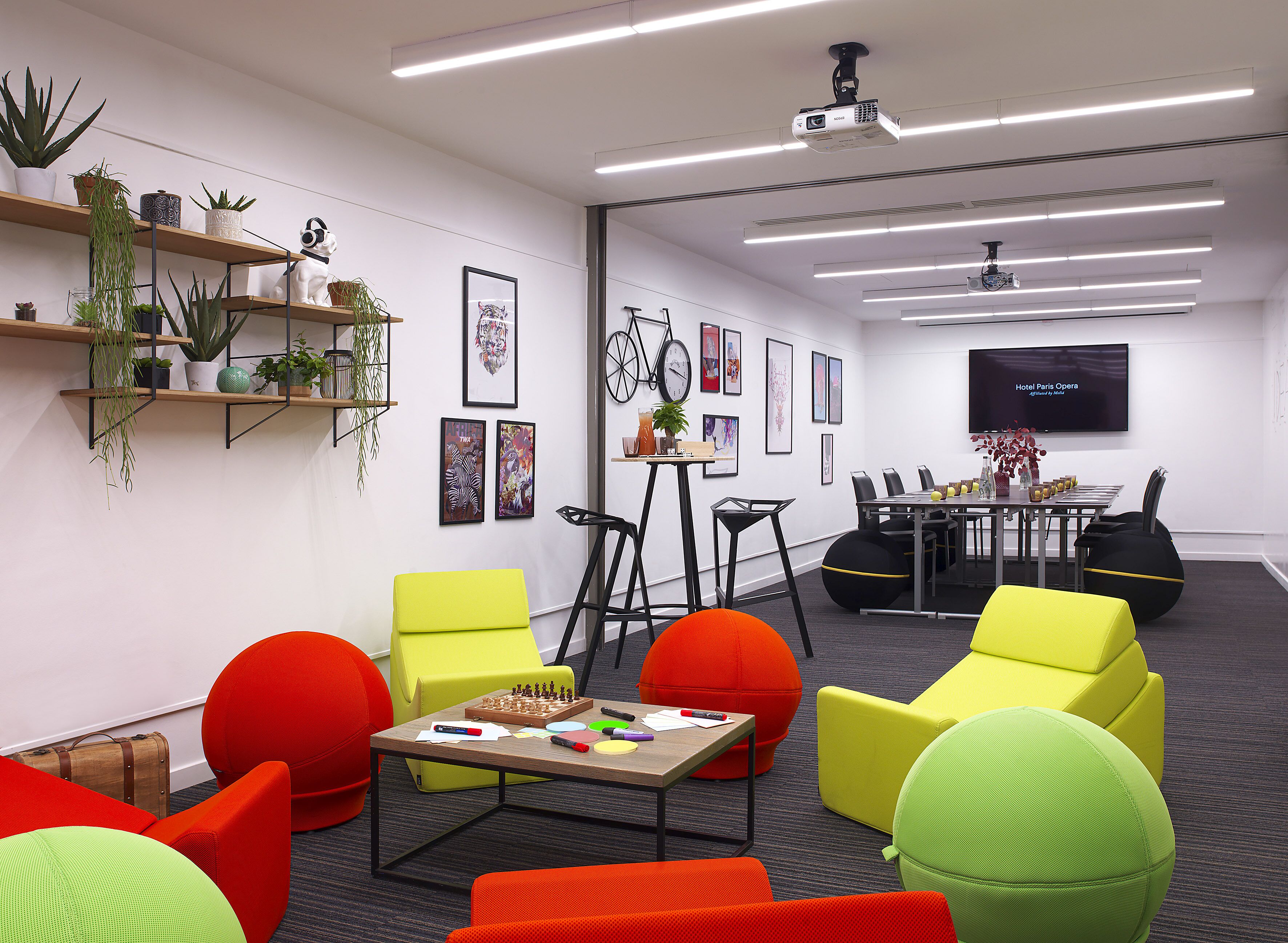 A modern meeting room with colorful chairs, a chessboard, and a large screen displaying 'Hotel Paris Opera'.